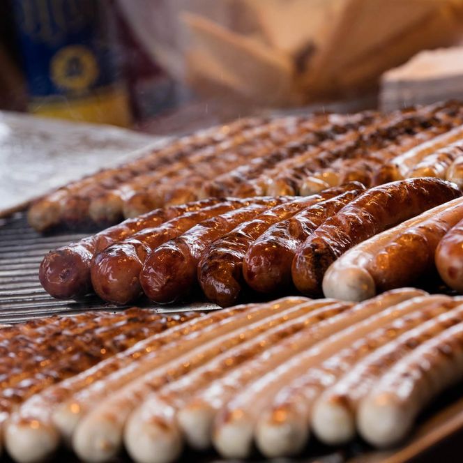 „Mehrere Reihen Bratwürste liegen dicht nebeneinander auf einem heißen Grill. Die Würste sind teils goldbraun gebraten, im Vordergrund ist ein Senfspender angeschnitten zu sehen.“
