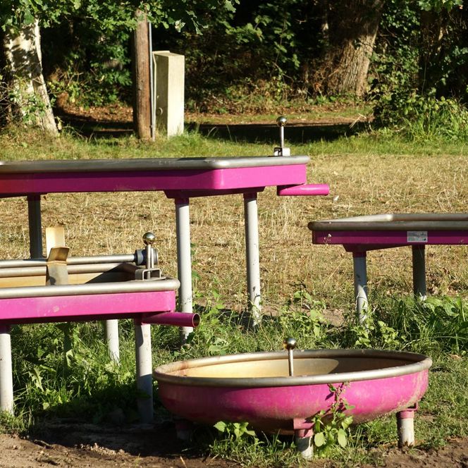 „Ein bunter Wasserspielplatz mit pinkfarbenen Wasserkanälen, Drehrädern und einem lustigen Wasserhahn in Form einer Figur mit Gesicht.“