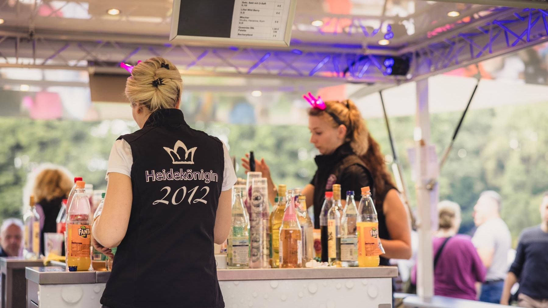 „Zwei Frauen bedienen an einem Getränkewagen. Die Frau im Vordergrund trägt ein schwarzes Shirt mit der Aufschrift ‚Heideblütenfest 2022‘ auf dem Rücken. Im Hintergrund stehen Menschen an der Theke.“