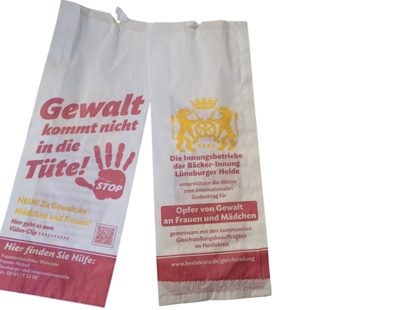 Zwei weiße Papiertüten mit roter und gelber Aufschrift. Die linke Tüte zeigt in roter Schrift den Text 'Gewalt kommt nicht in die Tüte!' mit einem roten Handabdruck und dem Wort 'STOP'. Darunter steht in kleinerer Schrift 'NEIN! Zu Gewalt an Mädchen und Frauen!' und 'Hier finden Sie Hilfe:' gefolgt von Kontaktinformationen. Die rechte Tüte trägt ein gelbes Wappen mit zwei Löwen und einer Krone sowie den Text 'Die Innungsbetriebe der Bäcker-Innung Lüneburger Heide unterstützen die Aktion zum internationalen Gedenktag für Opfer von Gewalt an Frauen und Mädchen'. Darunter steht in roter Schrift 'Gemeinsam mit den kommunalen Gleichstellungsbeauftragten im Heidekreis' und eine Webadresse.