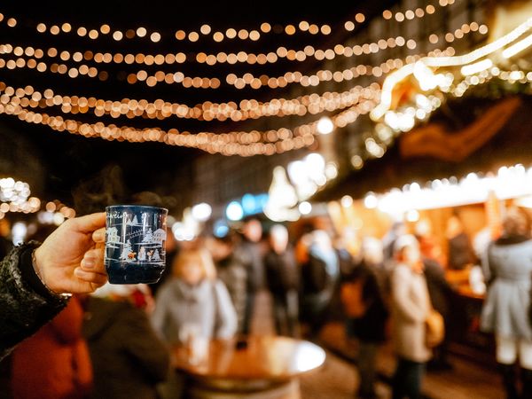 Hand, die eine Tasse Glühwein hält im Fokus und das beleuchtete Einbecker Weihnachtsdorf dahinter.