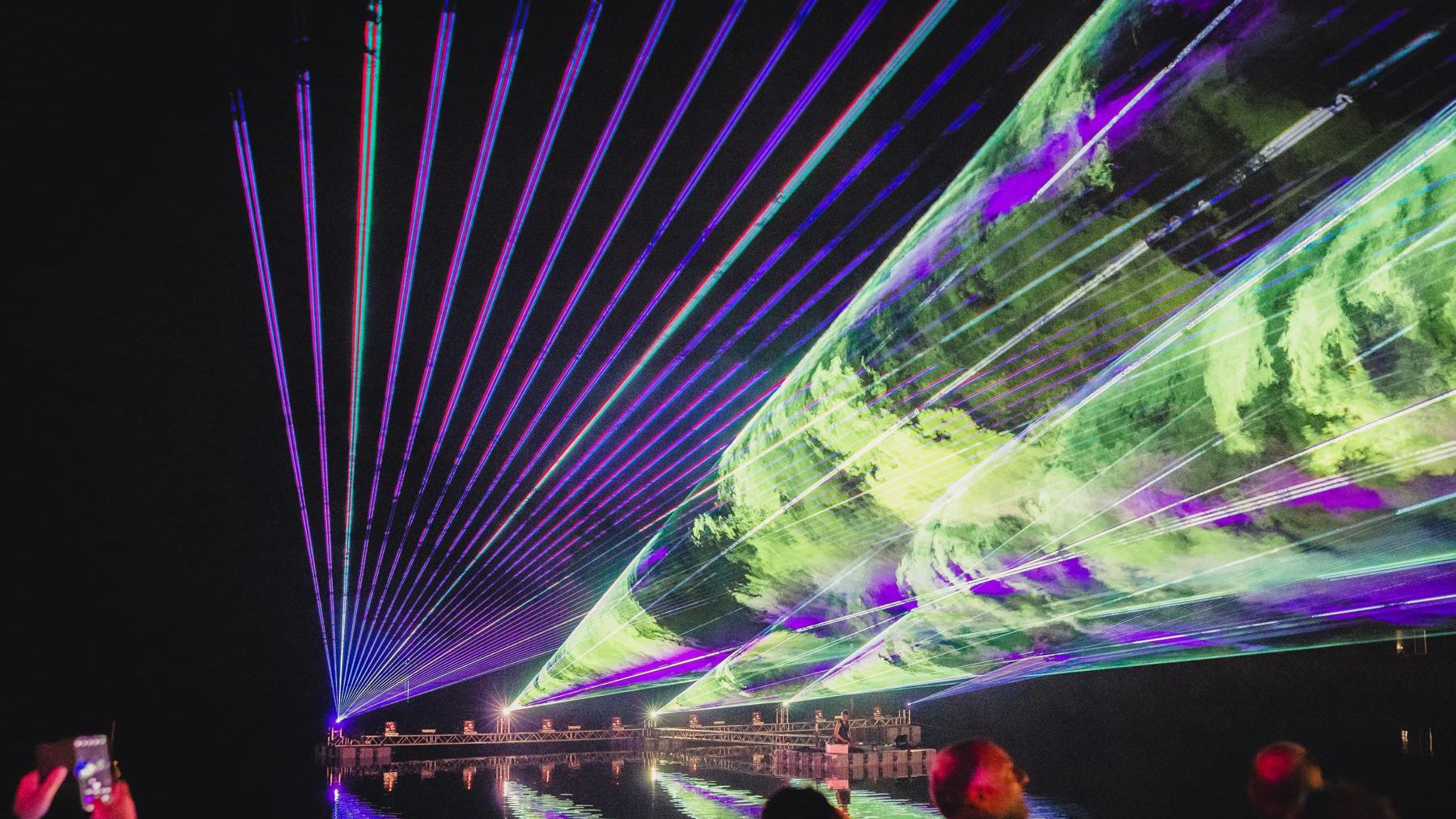 „Bunte Lasereffekte in Lila, Grün und Blau durchziehen den Nachthimmel. Im Vordergrund stehen Zuschauer mit leuchtenden Knicklichtern und Handys, die das Spektakel filmen.“