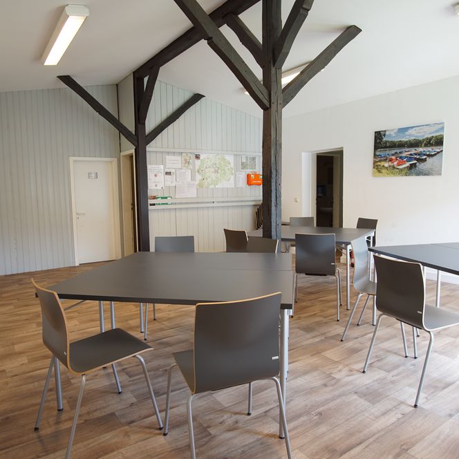 Ein heller Raum mit modernen Tischen und Stühlen, Holzboden und sichtbarem Deckenbalken.