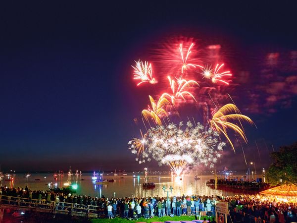 Farbenprächtiges Feuerwerk erleuchtet den Nachthimmel über einem Festival am Ufer eines Sees.