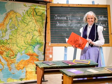 Eine Lehrerin steht in einem historischen Klassenraum in der Ausstellung "Von Federkiel und Rechenschieber"