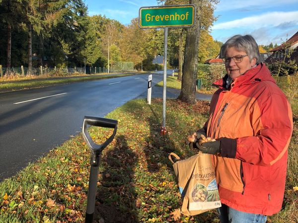 Person in oranger Jacke und blauer Jeans steht neben einem Straßenbegrenzungspfahl und einem grünen Ortsschild mit der Aufschrift 'Grevenhof'. Im Vordergrund steht ein Spaten im Boden.