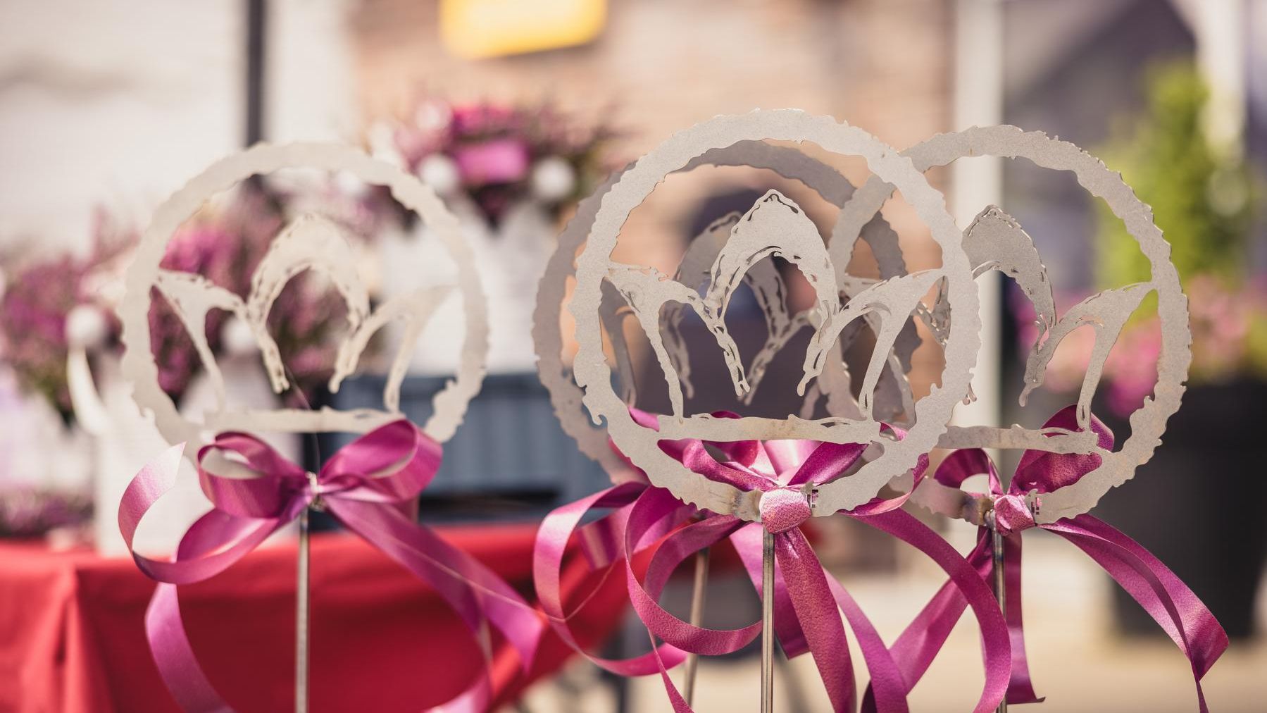 „Mehrere weiße Stäbe mit ausgeschnittenen Kronenmotiven und pinken Schleifen stehen in einer Reihe. Im Hintergrund ist ein roter Tisch mit Blumenschmuck leicht verschwommen zu sehen.“