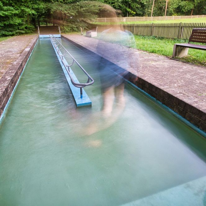 „Eine Person geht barfuß durch ein Wasserbecken einer Kneippanlage. Die Bewegung ist durch lange Belichtungszeit verschwommen dargestellt. Links und rechts stehen Bänke, im Hintergrund sind Bäume und ein Zaun zu sehen.“