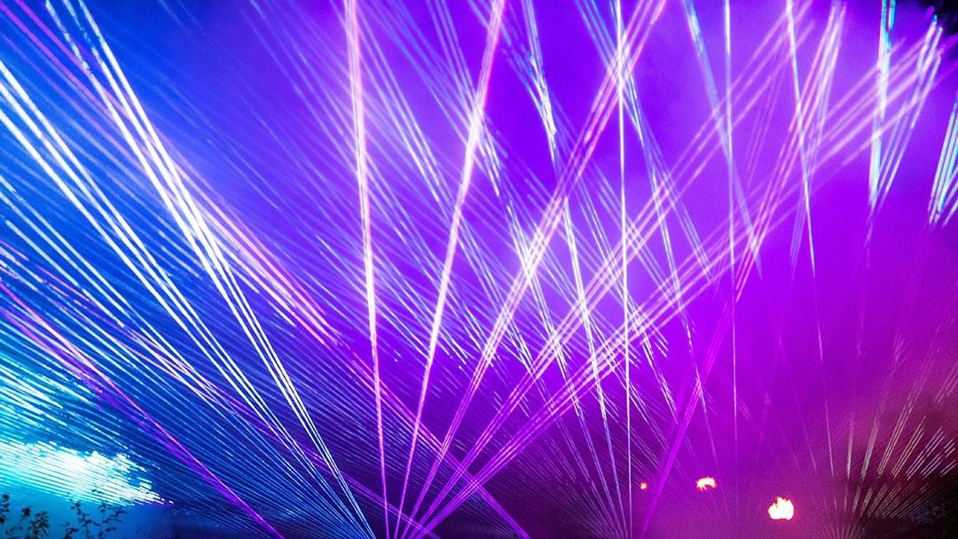 „Farbenfrohe Lichtstrahlen in Blau, Pink und Lila erleuchten die Nacht bei einer Lichtershow. Im Vordergrund sind dunkle Silhouetten von Menschen zu erkennen.“