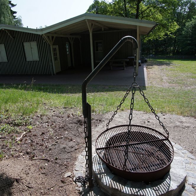 Runde Feuerstelle mit Grillrost und Kette vor einem überdachten Holzgebäude im Waldgebiet.