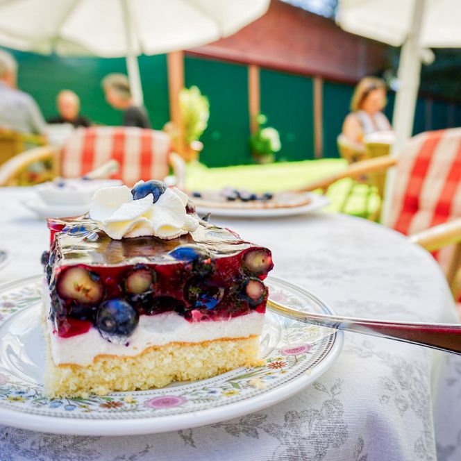 „Ein Stück Beerentorte mit Sahnetupfer steht auf einem verzierten Porzellanteller. Im Hintergrund sitzen Menschen auf gepolsterten Stühlen unter Sonnenschirmen im Garten eines Cafés.“