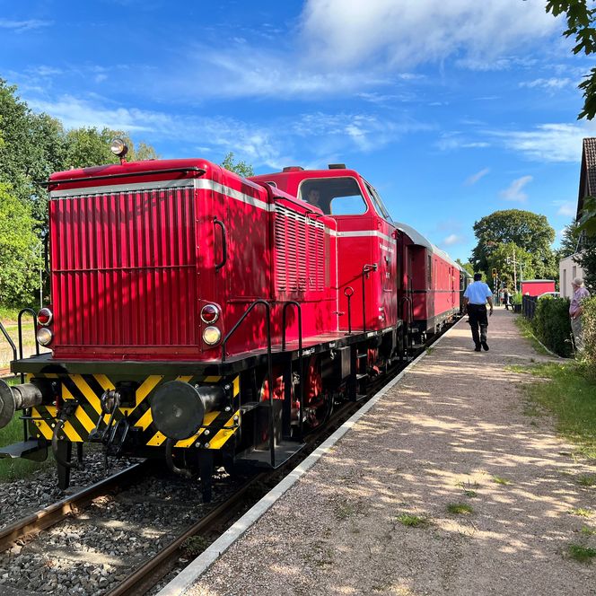 Rote Lokomotive am Bahnhofsgleis, daneben Schotterweg und Bäume, blauer Himmel mit einzelnen Wolken