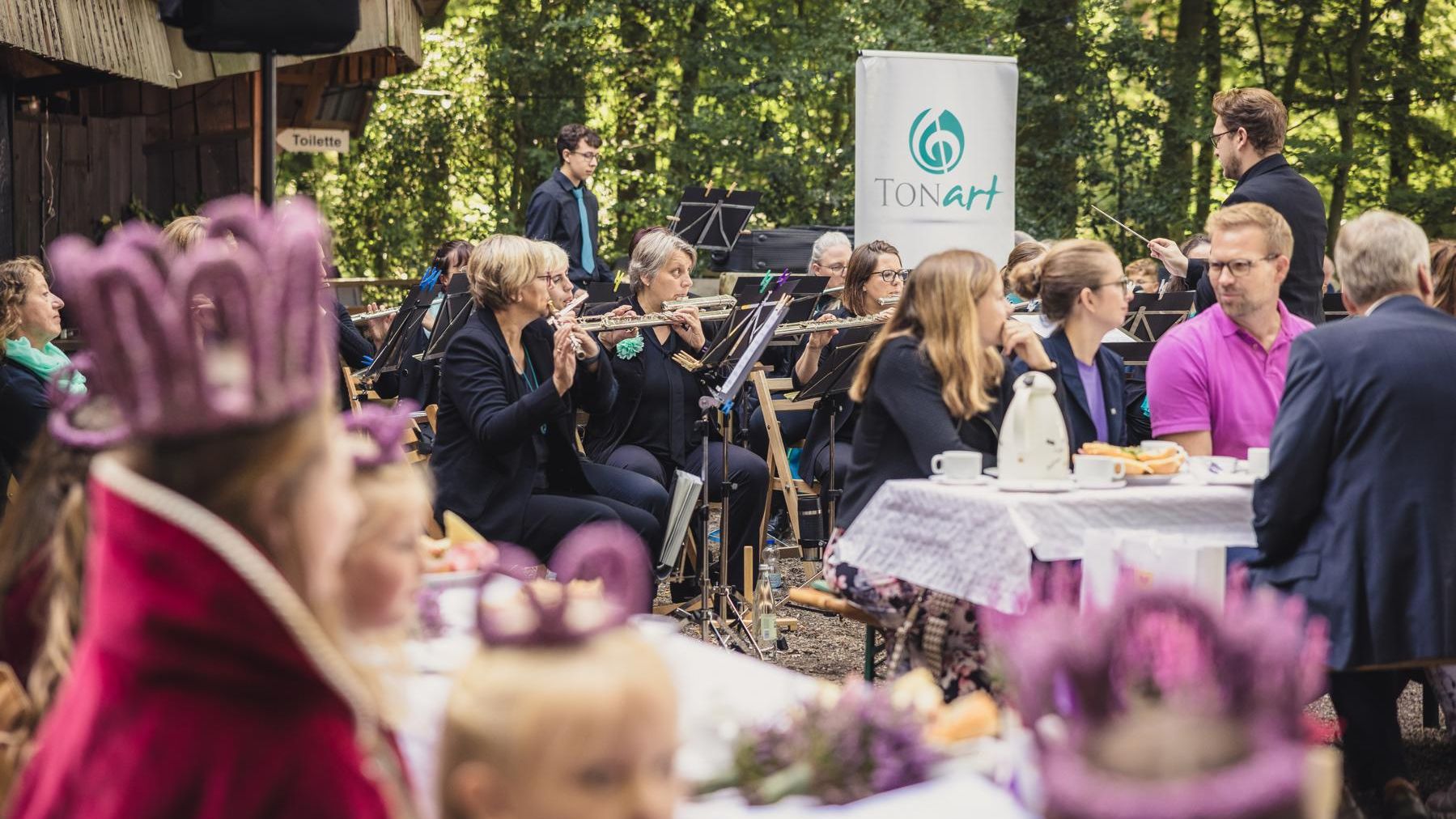 „Im Vordergrund sind Mädchen mit lilafarbenen Kronen zu sehen. Im Hintergrund sitzen Menschen an weiß gedeckten Tischen im Freien, während ein Blasorchester spielt. Dahinter hängt ein Banner der Samtgemeinde Amelinghausen.“
