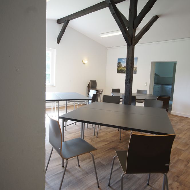 Ein heller Raum mit modernen Tischen und Stühlen, Holzboden und sichtbarem Deckenbalken.