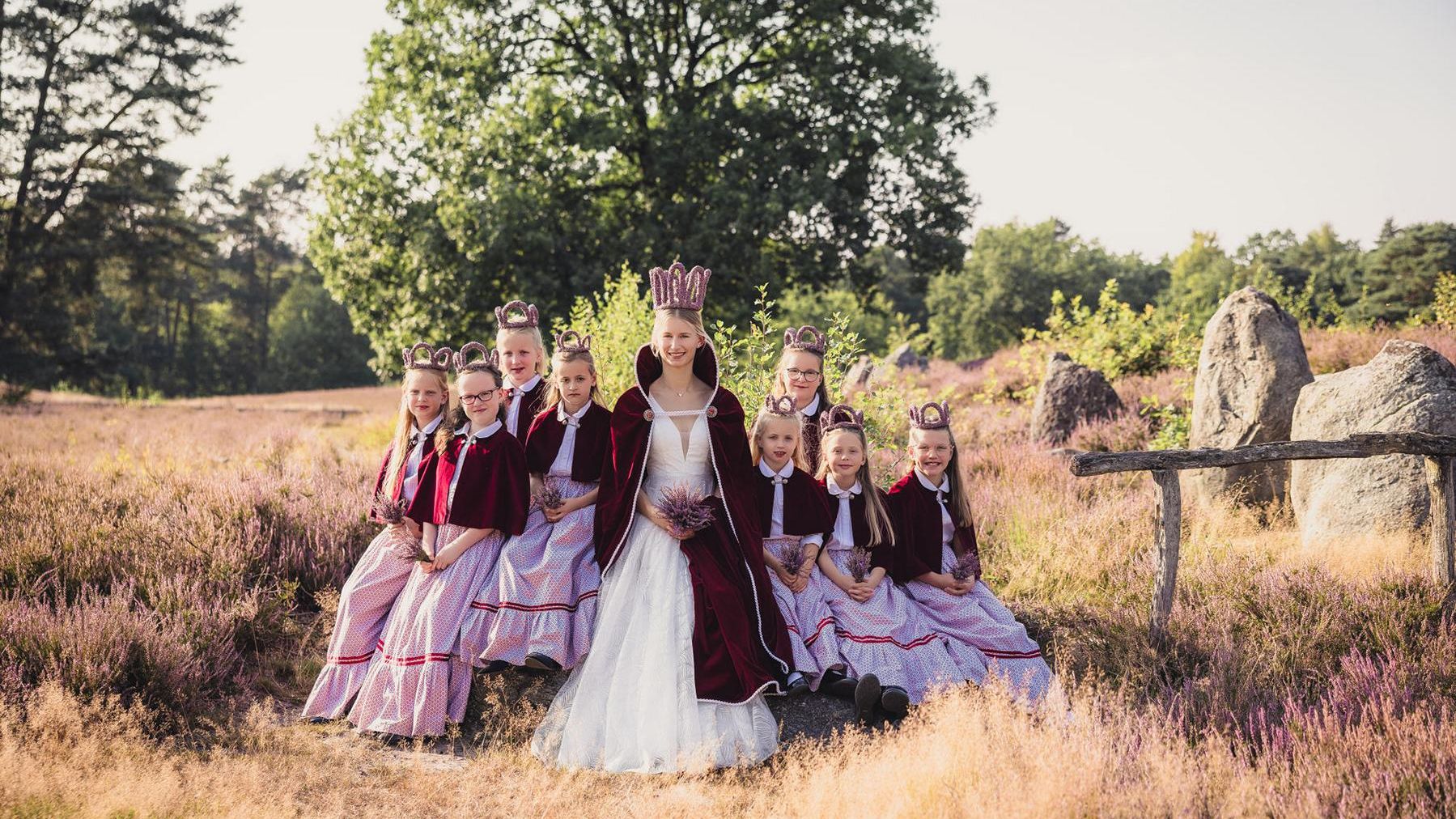 „Junge Frau in festlicher Robe mit Krone (Heidekönigin) sitzt inmitten einer Gruppe von Hofdamen in traditioneller Kleidung. Die Gruppe posiert in der Heide bei sommerlichem Wetter.“