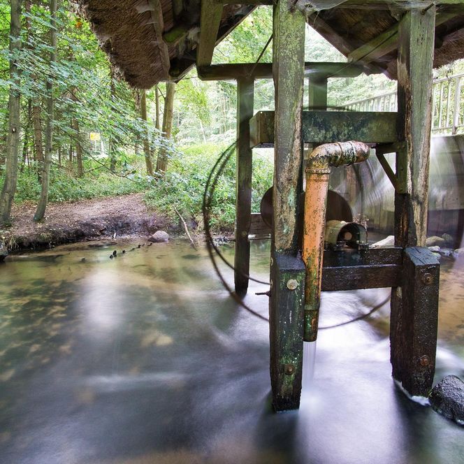 „Ein altes, drehendes Wasserrad unter einem kleinen Holzdach an einem klaren Bachlauf im Wald.“