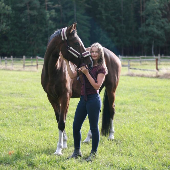 „Eine junge Frau steht auf einer grünen Wiese neben einem braunen Pferd mit weißer Blesse. Sie hält das Pferd am Halfter und schaut in die Kamera. Im Hintergrund sind ein Holzzaun und Bäume zu sehen.“