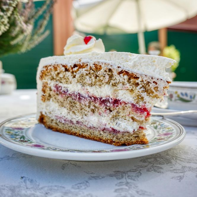 „Ein großes Stück Buchweizentorte mit Sahnefüllung steht auf einem Teller auf einem gedeckten Gartentisch. Im Hintergrund sind Gäste, Teetassen und Sonnenschirme zu sehen.“